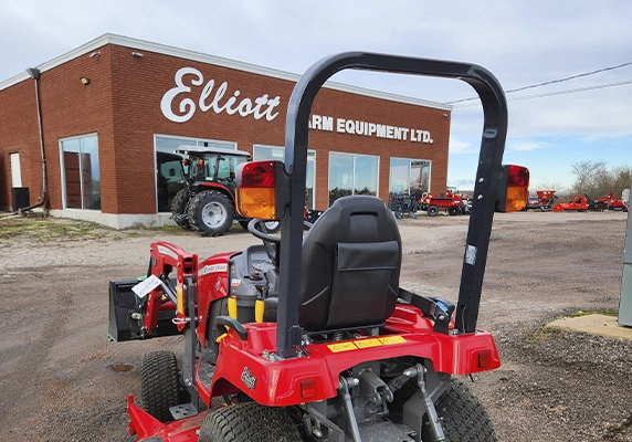 Compact red utility tractor parked outside Elliott Farm Equipment dealership building, with larger tractors visible in the background.